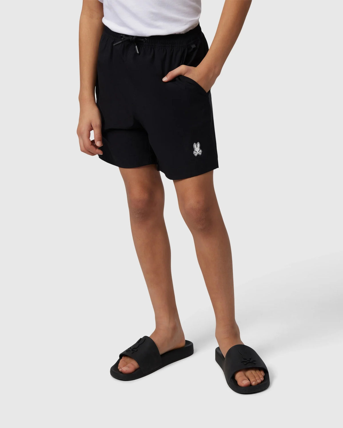 A person stands displaying Psycho Bunny hydrochromic swim trunks and black slide sandals, the trunks feature a small white logo on the left thigh. Only the lower half of the person is visible against a plain background.