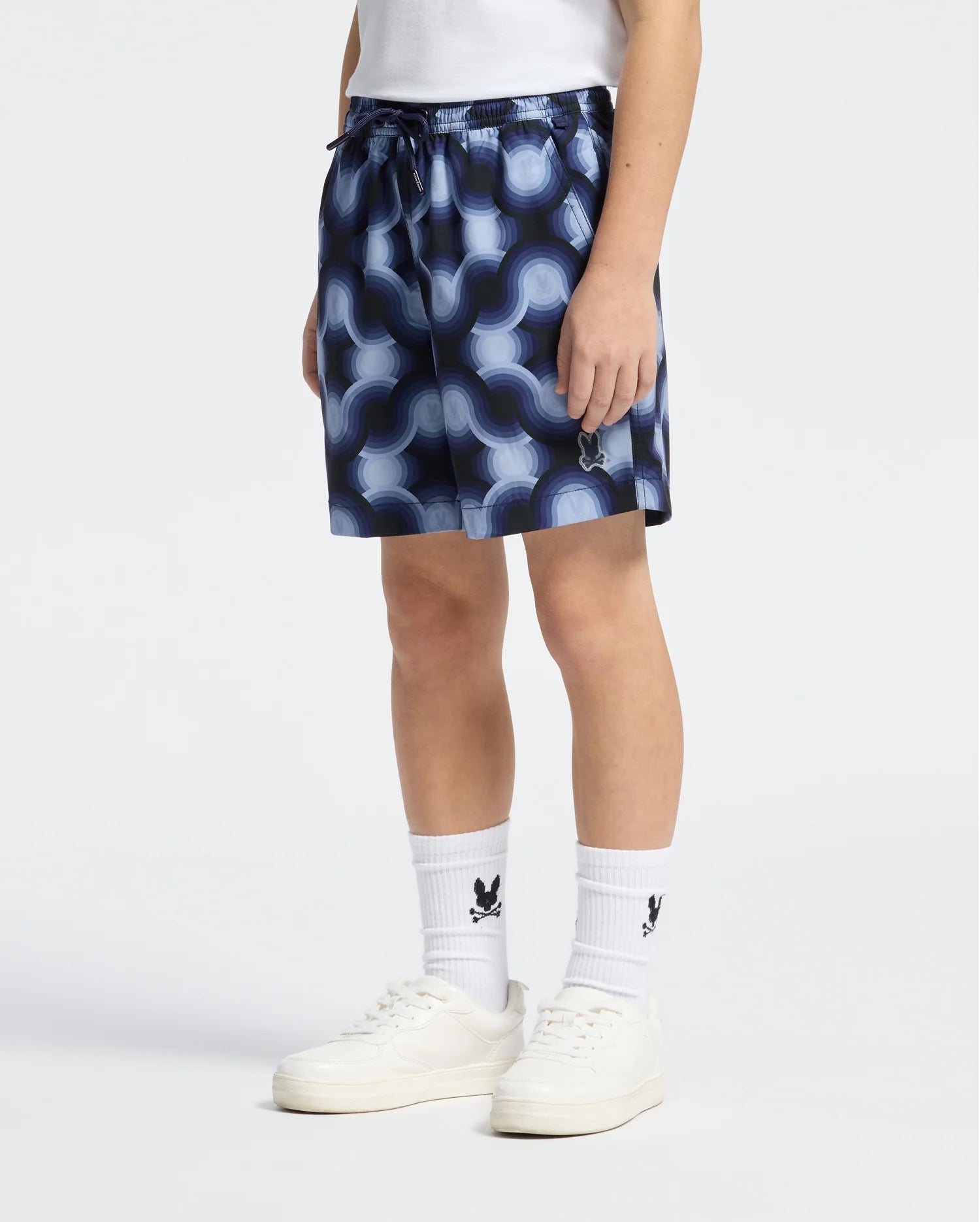 A person in a white shirt and KIDS PAROS PRINTED SWIM TRUNK - B0W450F200 by Psycho Bunny stands against a plain backdrop, wearing white logo socks and sleek white sneakers, exuding casual style.
