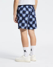 Wearing Psycho Bunny's KIDS PAROS PRINTED SWIM TRUNK - B0W450F200, along with white sneakers and black-logo socks, a person stands against a plain backdrop.