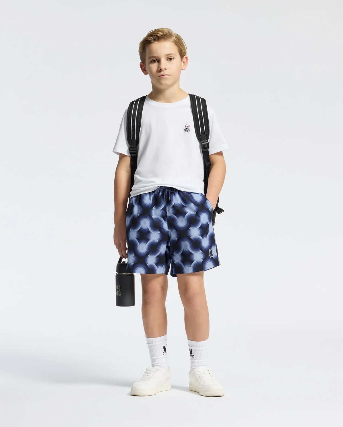 A young boy poses against a plain backdrop in a white tee, wearing Psycho Bunny's KIDS PAROS PRINTED SWIM TRUNK - B0W450F200. He's paired them with white socks and sneakers, slings a waterproof bag over his shoulder, and holds a black bottle; his expression is neutral.
