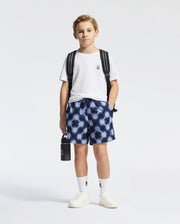 A young boy poses against a plain backdrop in a white tee, wearing Psycho Bunny's KIDS PAROS PRINTED SWIM TRUNK - B0W450F200. He's paired them with white socks and sneakers, slings a waterproof bag over his shoulder, and holds a black bottle; his expression is neutral.