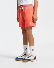 A person is wearing Psycho Bunny's KIDS SAMOS HYDROCHROMIC SWIM TRUNK featuring a rabbit logo, white socks with the same emblem, a white shirt, and blue-and-white sneakers. The quick-dry fabric offers comfort while the plain white background showcases the vibrant coral outfit.