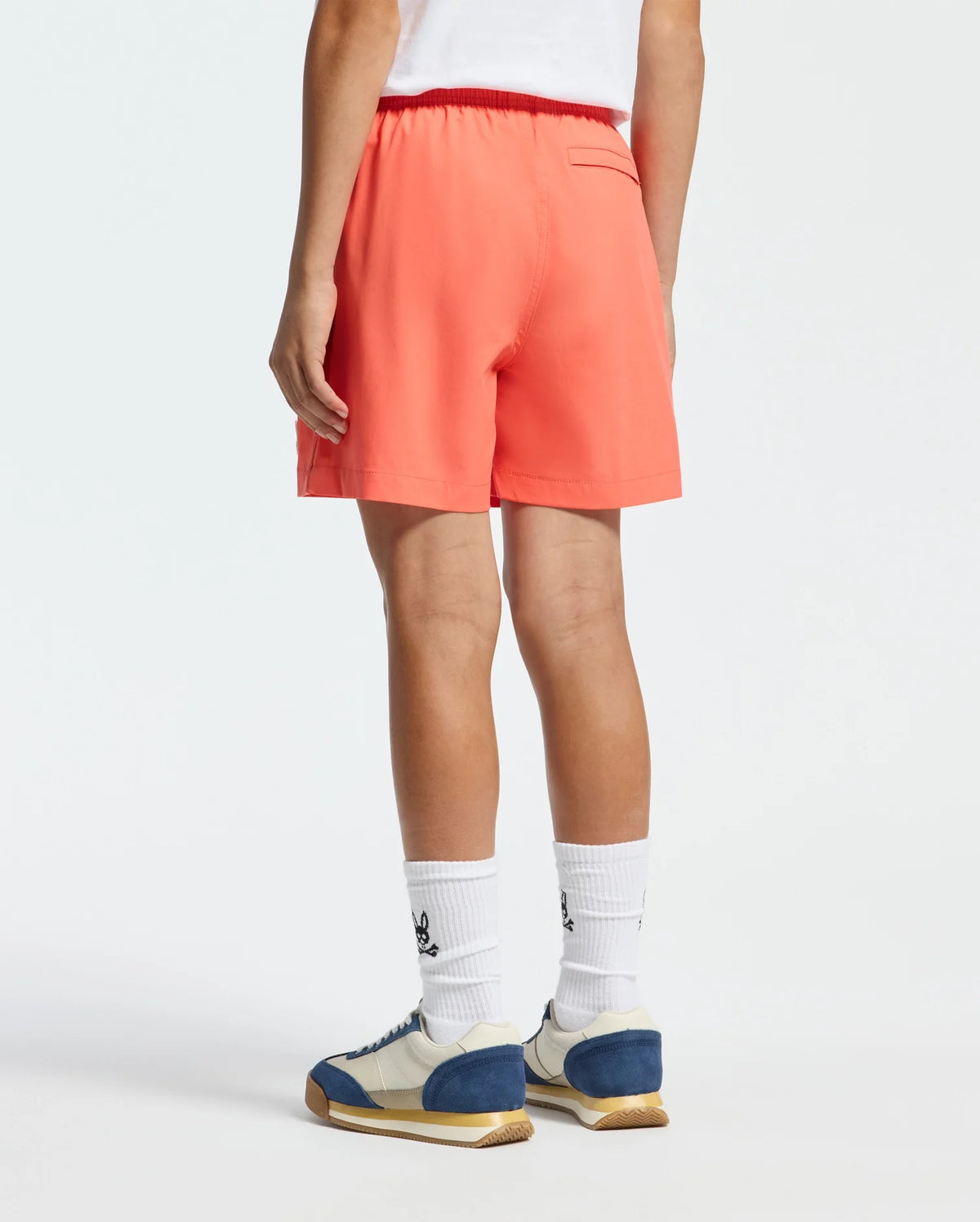 A person in Psycho Bunny's coral KIDS SAMOS HYDROCHROMIC SWIM TRUNK, featuring a back pocket, stands on a plain background with white socks sporting a design and white and blue sneakers.