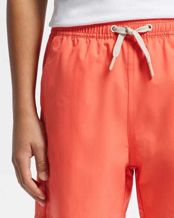A person wears Psycho Bunny's KIDS SAMOS HYDROCHROMIC SWIM TRUNK - B0W453F200, coral-colored and quick-dry with a drawstring. Their hand rests by the elastic waistband while a white shirt is seen above.