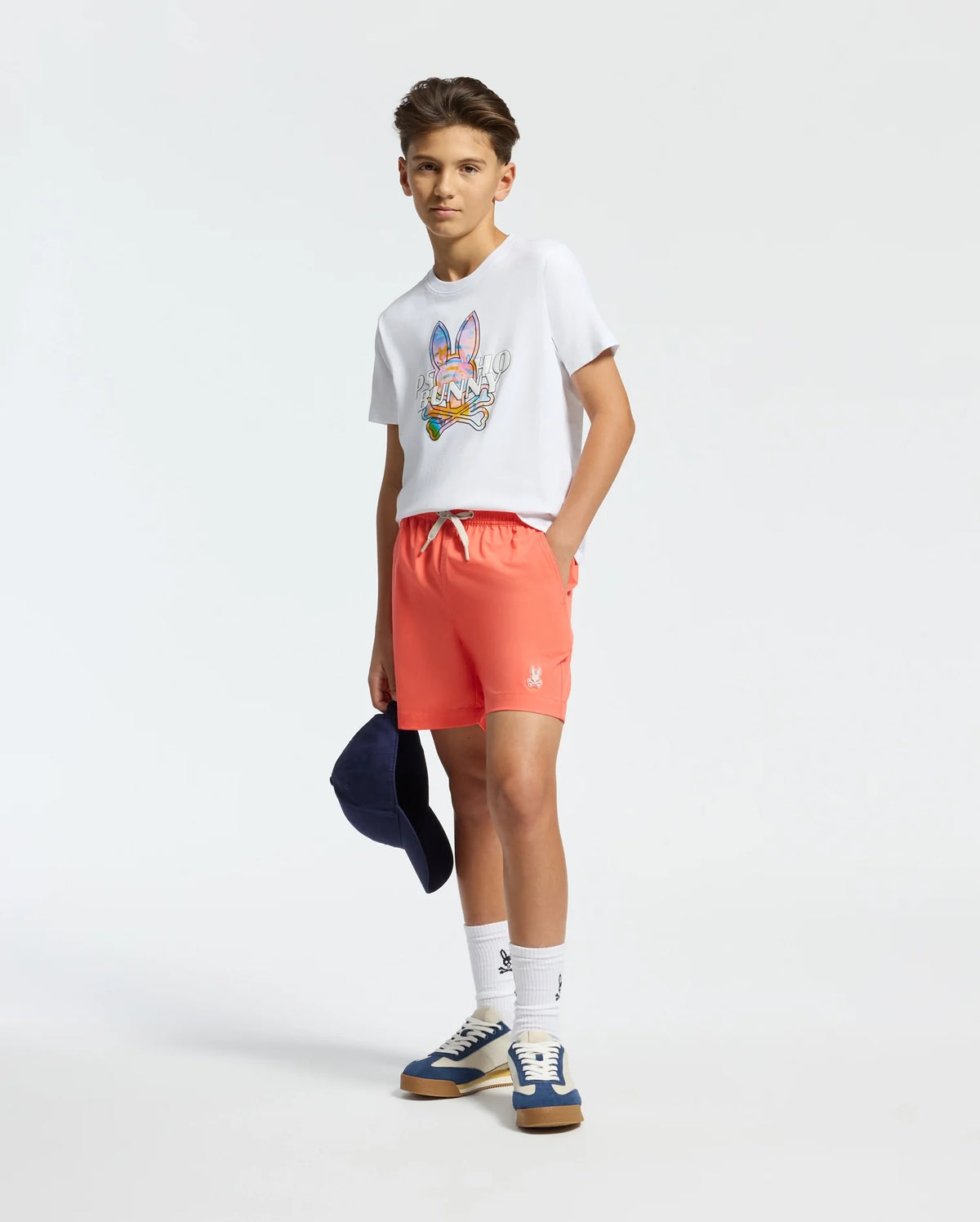 A teenage boy confidently wears a white tee with a colorful graphic, Psycho Bunny's KIDS SAMOS Hydrochromic Swim Trunks, and blue sneakers. He's holding a navy cap and wearing quick-dry white socks against a plain white background.