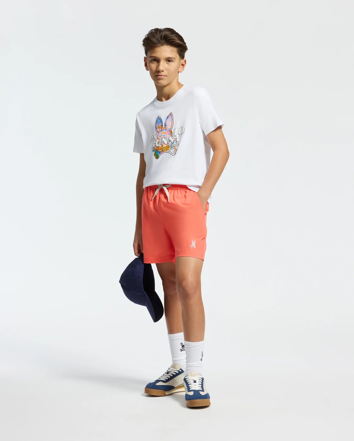A teenage boy confidently wears a white tee with a colorful graphic, Psycho Bunny's KIDS SAMOS Hydrochromic Swim Trunks, and blue sneakers. He's holding a navy cap and wearing quick-dry white socks against a plain white background.