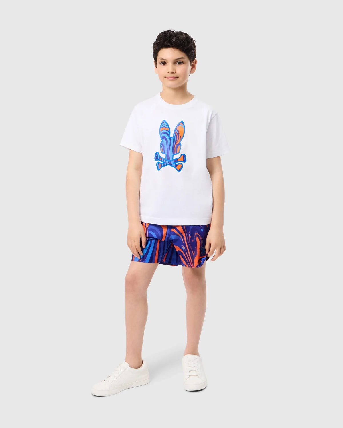 A young boy stands on a light grey background wearing a white T-shirt with a colorful abstract design. He sports vibrant blue and orange KIDS NEVADA ALL OVER PRINT SWIM TRUNK - B0W526C200 from Psycho Bunny, made from quick-dry fabric. With short dark hair and white sneakers, he gazes confidently at the camera with a relaxed posture.