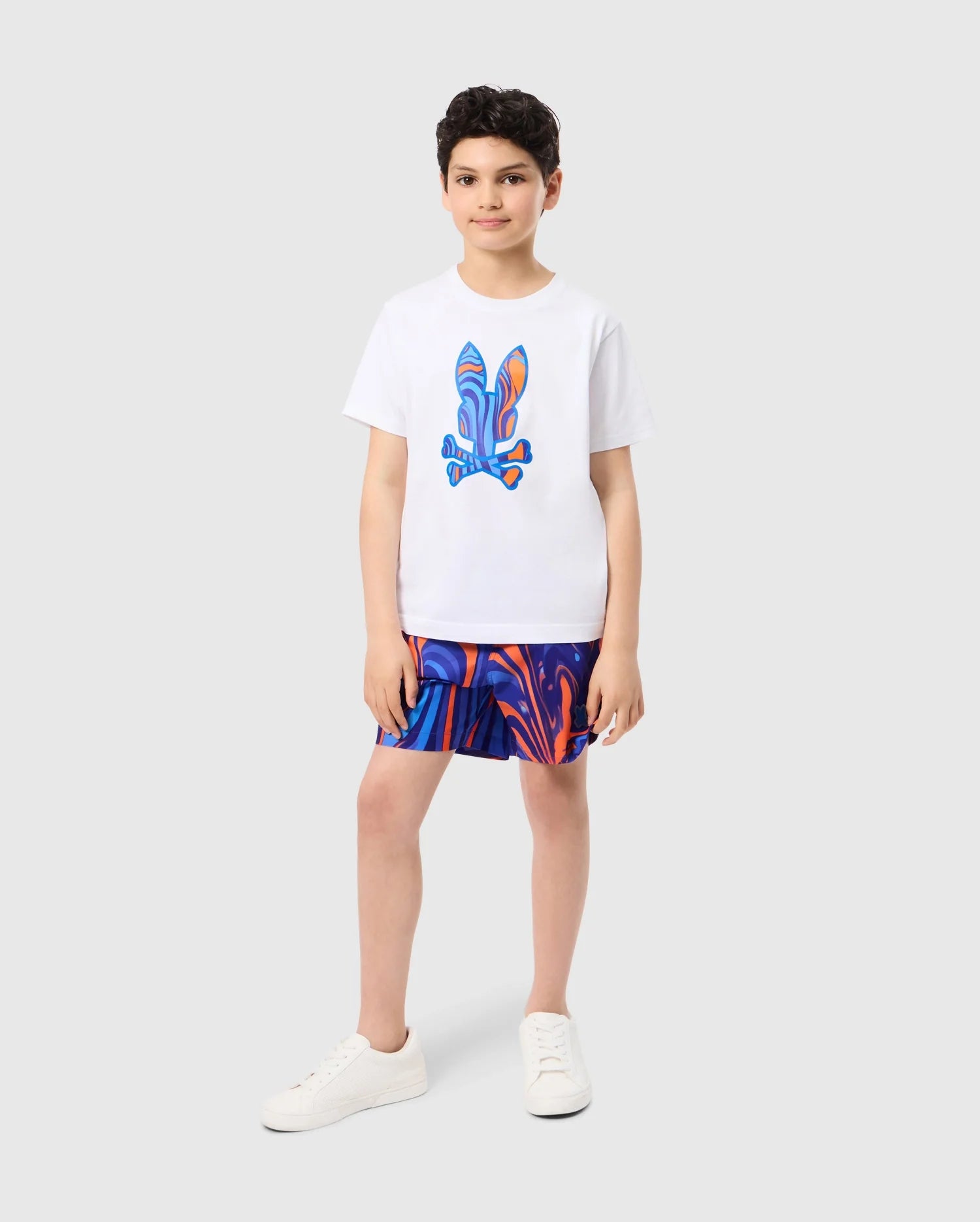 A young boy stands on a light grey background wearing a white T-shirt with a colorful abstract design. He sports vibrant blue and orange KIDS NEVADA ALL OVER PRINT SWIM TRUNK - B0W526C200 from Psycho Bunny, made from quick-dry fabric. With short dark hair and white sneakers, he gazes confidently at the camera with a relaxed posture.