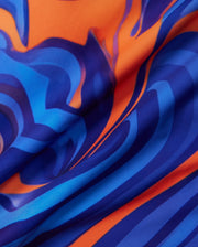 Close-up of the KIDS NEVADA ALL OVER PRINT SWIM TRUNK (B0W526C200) by Psycho Bunny, showcasing a vibrant swirling pattern in bold shades of blue and orange. The design appears fluid and dynamic, resembling abstract waves or flames. This quick-dry fabric has a smooth texture with folds that catch light, adding depth to the visual effect.