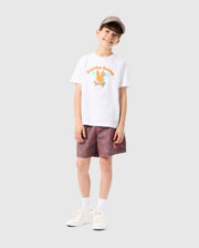 A smiling young boy stands against a plain background, wearing a white t-shirt with colorful graphics, KIDS SHELDON ALL OVER PRINT SWIM TRUNK - B0W588C200 from Psycho Bunny, white socks, and sneakers. He is also wearing a beige cap, and his hands are relaxed at his sides.
