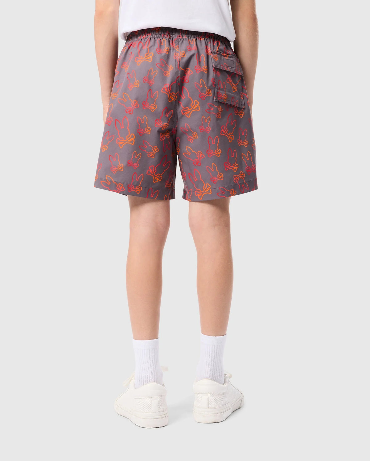 A person wearing gray KIDS SHELDON ALL OVER PRINT SWIM TRUNK - B0W588C200 by Psycho Bunny with a red bunny pattern is seen from behind. The shorts, made of quick-dry fabric, feature an elastic waistband and a back pocket. The individual also wears white socks, white sneakers, and a white t-shirt. The background is plain and light gray.