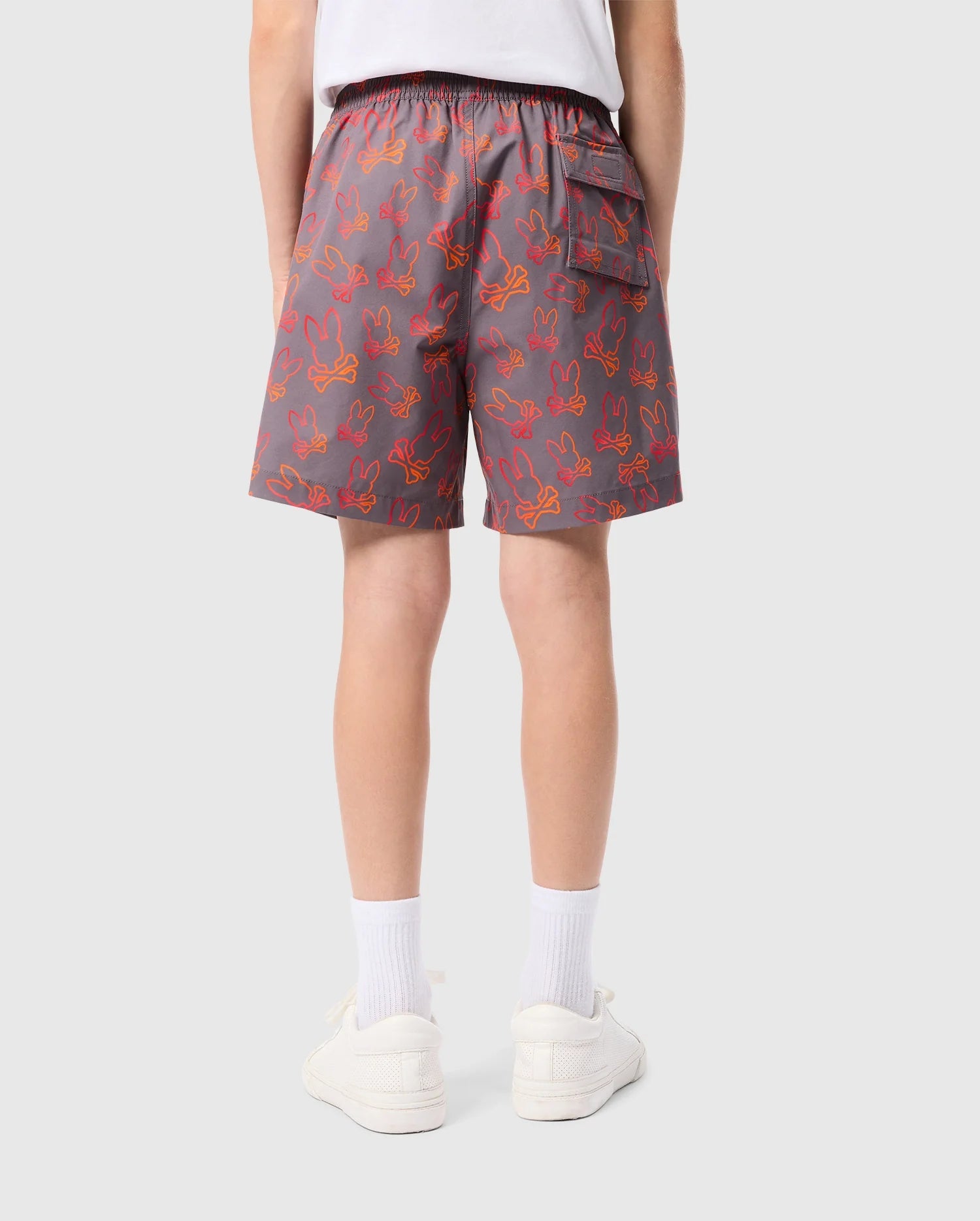 A person wearing gray KIDS SHELDON ALL OVER PRINT SWIM TRUNK - B0W588C200 by Psycho Bunny with a red bunny pattern is seen from behind. The shorts, made of quick-dry fabric, feature an elastic waistband and a back pocket. The individual also wears white socks, white sneakers, and a white t-shirt. The background is plain and light gray.