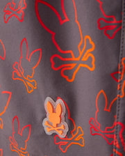 Close-up of Psycho Bunny KIDS SHELDON ALL OVER PRINT SWIM TRUNK - B0W588C200 featuring a bunny pattern with skulls and crossbones in red, orange, and yellow. There's a small rubberized patch of the same bunny skull and crossbones design in orange attached to the quick-dry fabric. The background is a muted gray color.