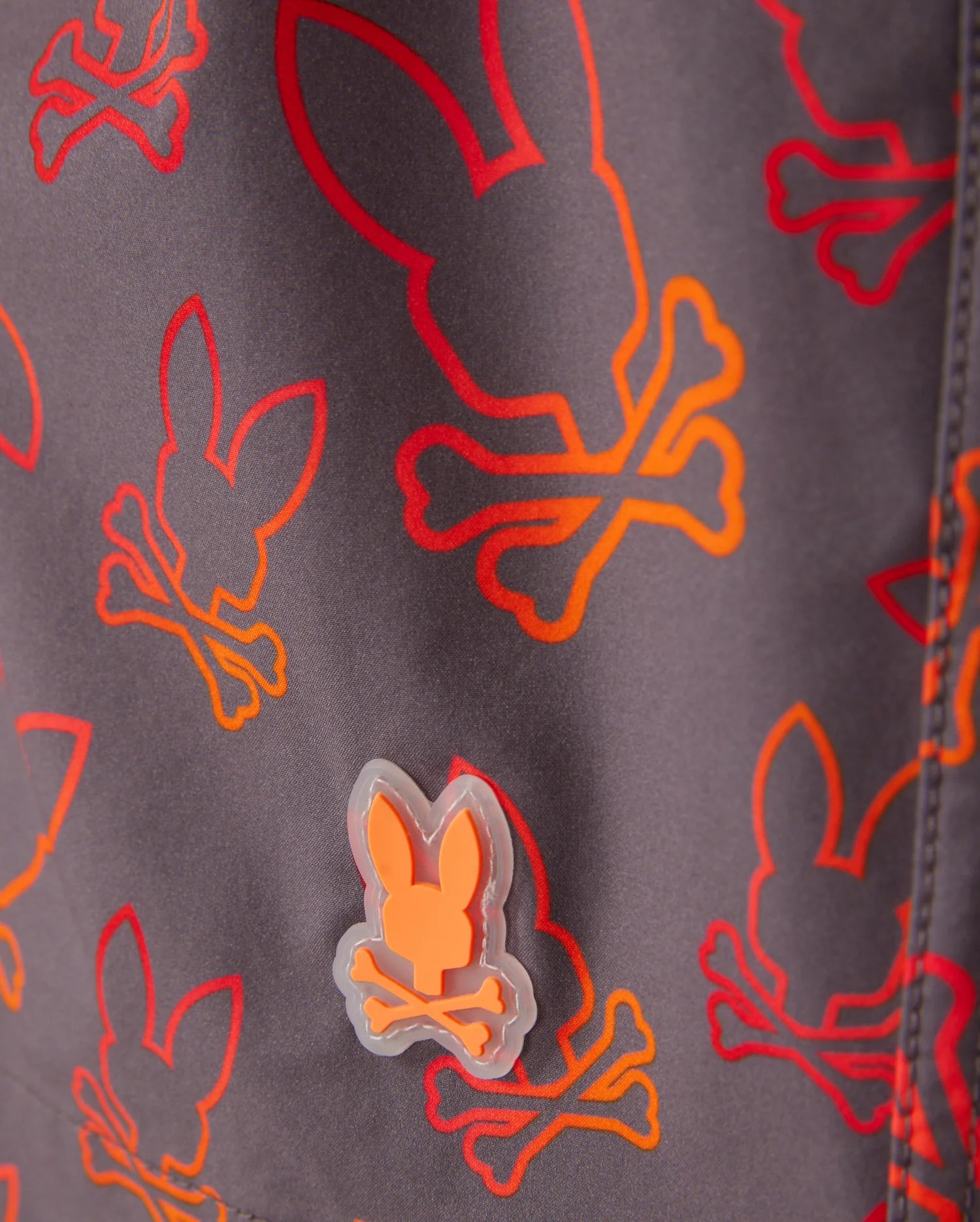 Close-up of Psycho Bunny KIDS SHELDON ALL OVER PRINT SWIM TRUNK - B0W588C200 featuring a bunny pattern with skulls and crossbones in red, orange, and yellow. There's a small rubberized patch of the same bunny skull and crossbones design in orange attached to the quick-dry fabric. The background is a muted gray color.