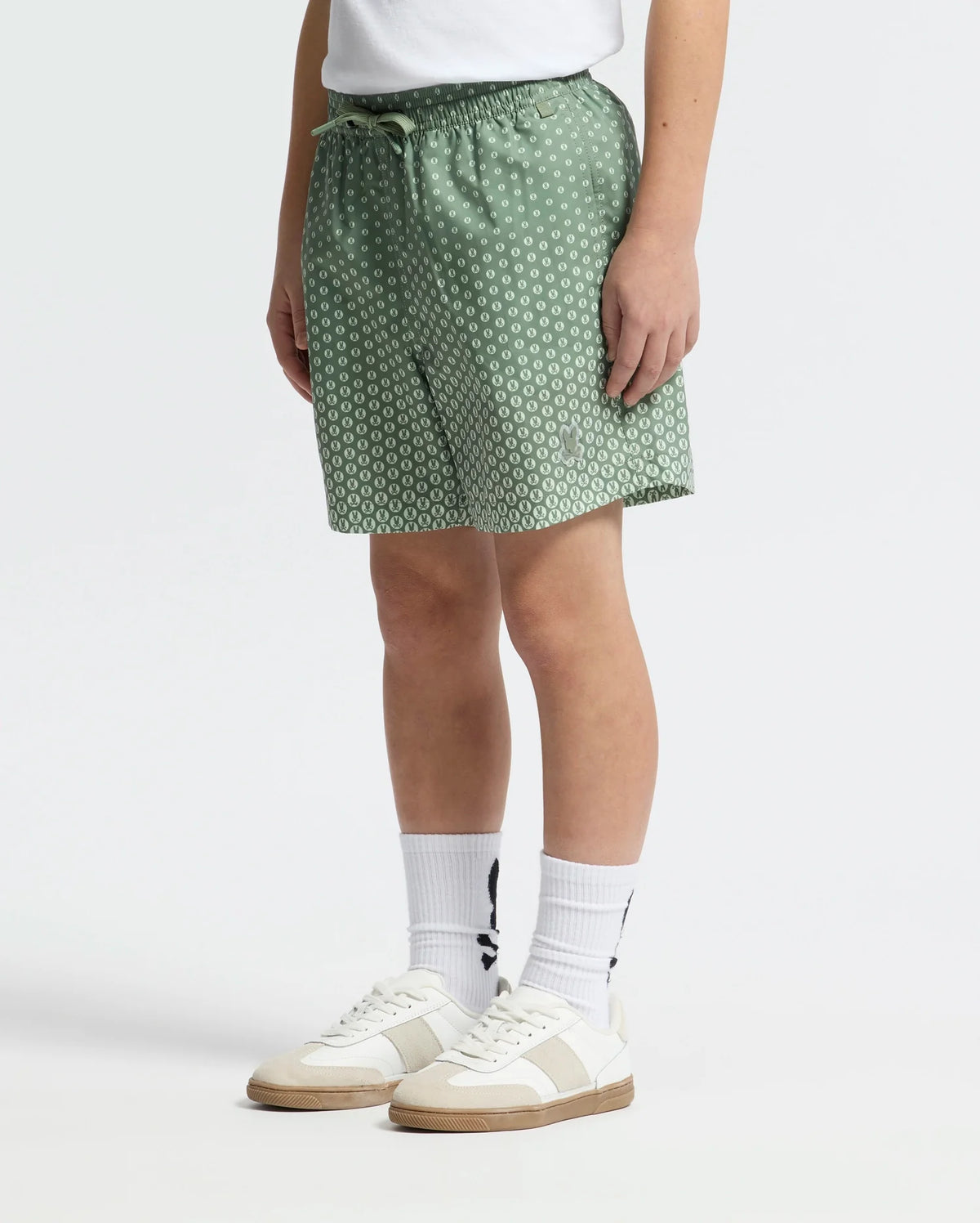 A person is wearing a white t-shirt, Psycho Bunny's KIDS BENDIGO PRINTED SWIM TRUNK in green, white socks with a subtle graphic, and white sneakers, standing against a plain background.