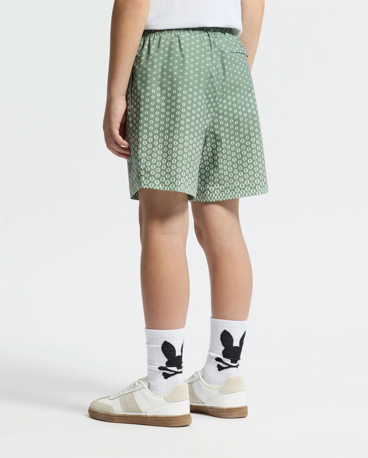 A person is shown waist down wearing Psycho Bunny's KIDS BENDIGO PRINTED SWIM TRUNK in green with a white geometric pattern, paired with white socks featuring a black design and white sneakers with beige accents. The background is plain white.