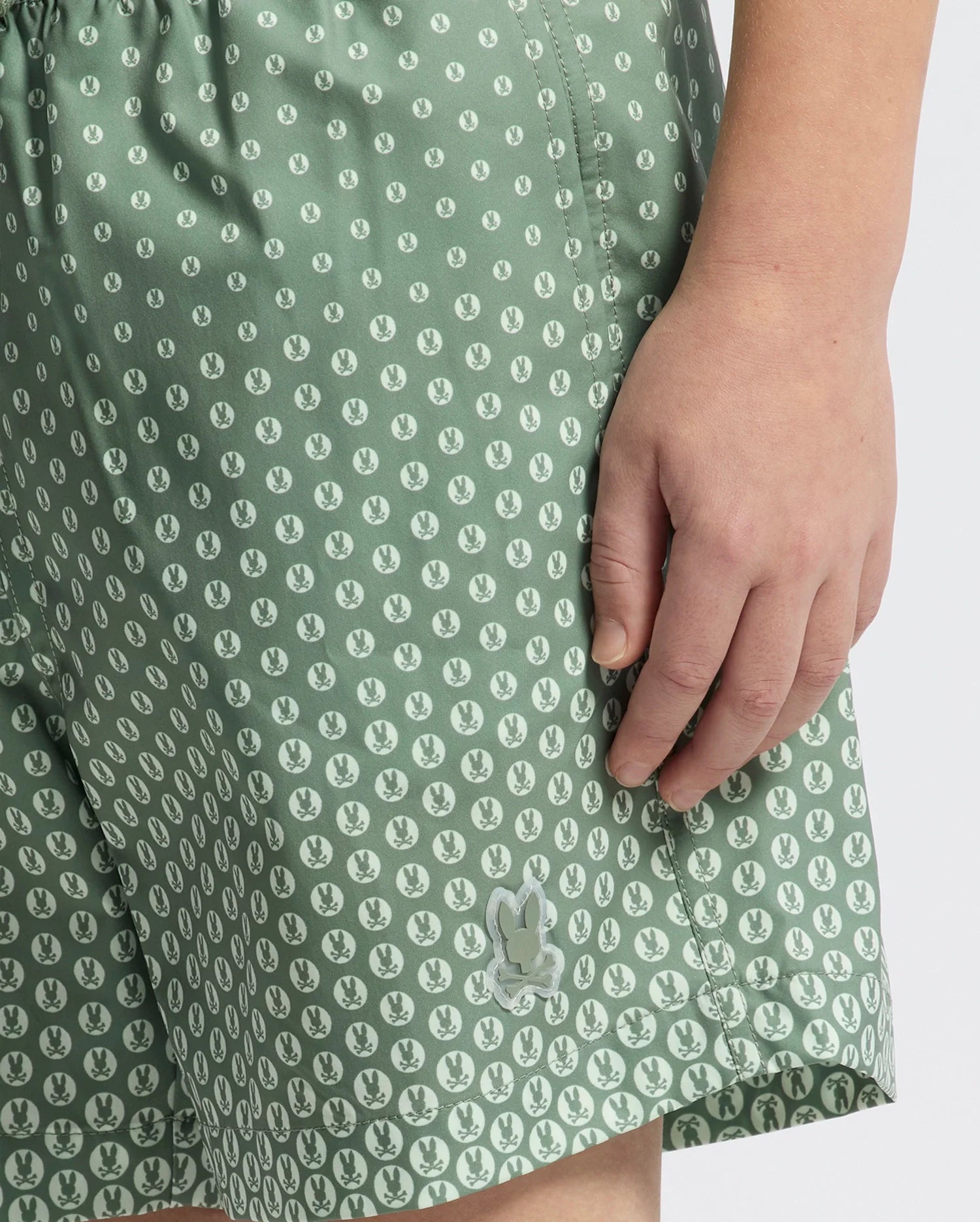Close-up of a person wearing KIDS BENDIGO PRINTED SWIM TRUNK by Psycho Bunny, featuring a bunny pattern and small white dots, crafted from quick-dry fabric. The person's right hand rests by their side.