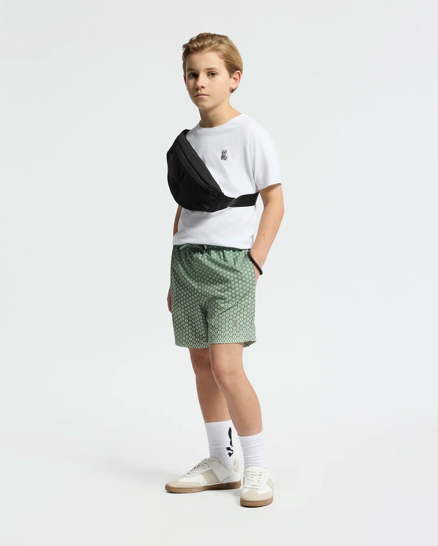 A young boy in green Psycho Bunny KIDS BENDIGO PRINTED SWIM TRUNK - B0W619F200 stands against a white background, wearing a white t-shirt, a black waterproof waist bag across his chest, and white sneakers with tall socks.