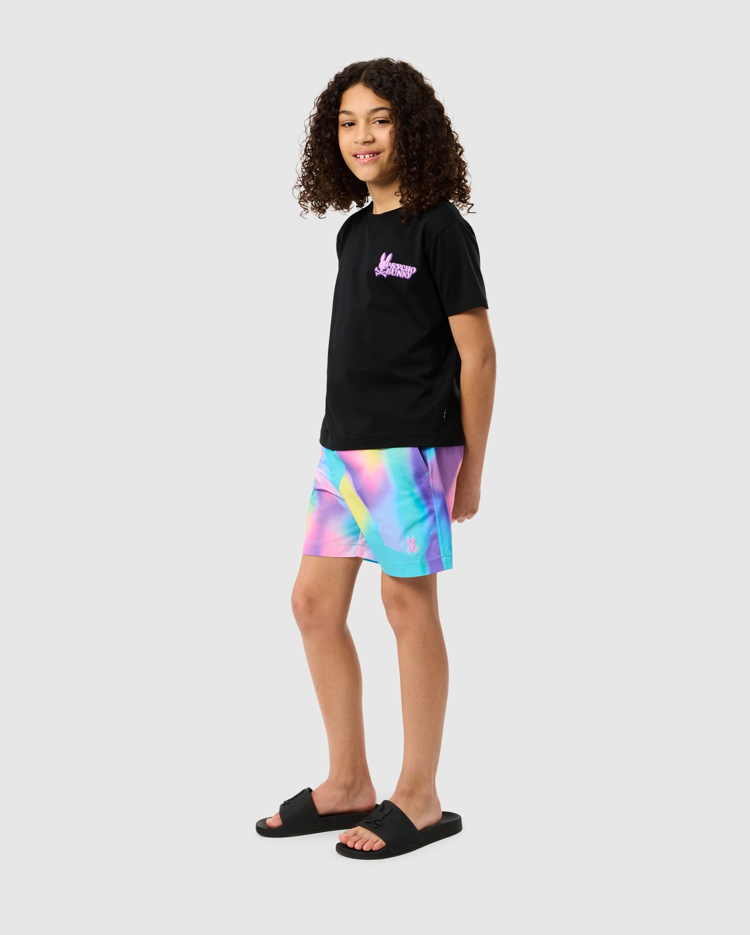 A young person with curly hair wearing a black t-shirt with a logo on the chest, Psycho Bunny KIDS TYLER PRINT SWIM TRUNK - B0W620C200 resembling boys' swimwear, and black slide sandals. They are standing against a plain gray background, looking to the side with a slight smile.