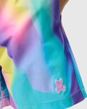A close-up of boys' swimwear featuring a vibrant, multicolor tie-dye design in shades of yellow, pink, purple, and blue. On the lower right side of these KIDS TYLER PRINT SWIM TRUNK - B0W620C200 by Psycho Bunny, there is a small icon of a bunny head with crossed bones underneath it.