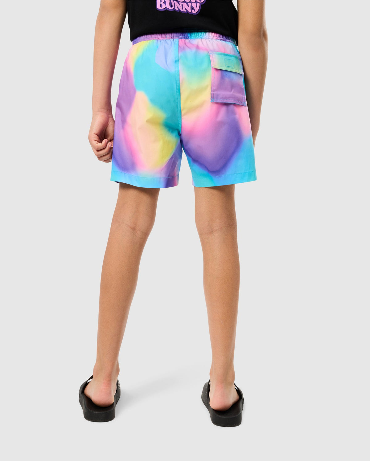 A child wearing vibrant, multicolored shorts featuring a bold tie-dye pattern and a black t-shirt is standing with their back to the camera. The KIDS TYLER PRINT SWIM TRUNK - B0W620C200 from Psycho Bunny showcases quick-dry fabric, while the t-shirt has a small graphic and text on the upper back. The child is also wearing black flip-flops.