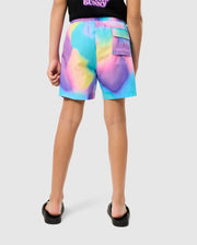 A child wearing vibrant, multicolored shorts featuring a bold tie-dye pattern and a black t-shirt is standing with their back to the camera. The KIDS TYLER PRINT SWIM TRUNK - B0W620C200 from Psycho Bunny showcases quick-dry fabric, while the t-shirt has a small graphic and text on the upper back. The child is also wearing black flip-flops.