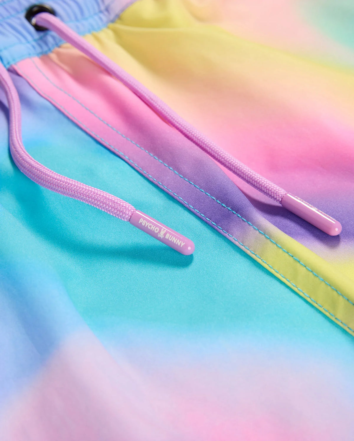 Close-up of pastel-colored fabric featuring a gradient of blue, purple, pink, and yellow hues. The drawstring on this KIDS TYLER PRINT SWIM TRUNK - B0W620C200 is light purple with plastic tips, one labeled "Psycho Bunny." The stitching along the edge highlights its high-quality quick-dry fabric and multicolor design.
