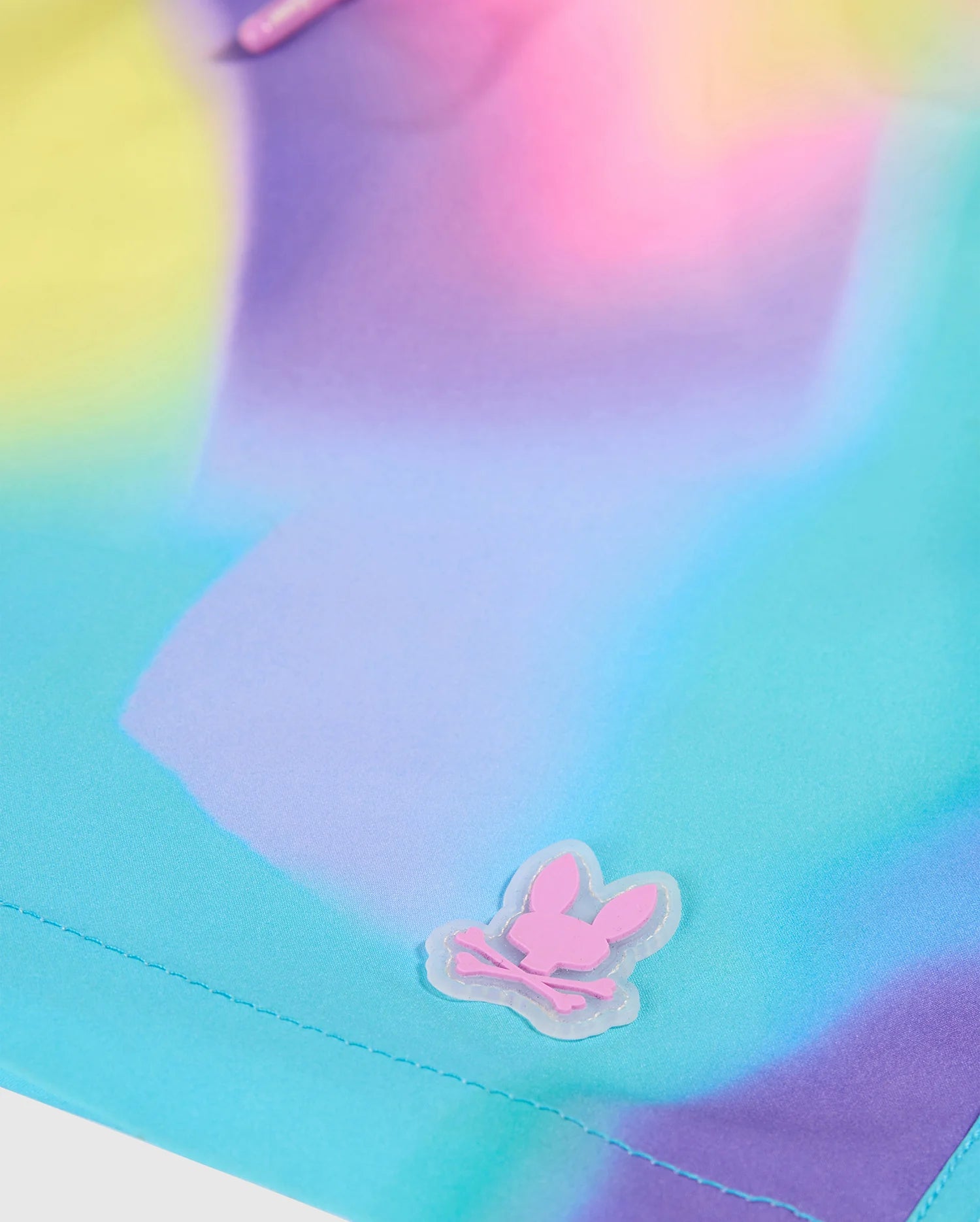 A close-up of a pastel tie-dye fabric with shades of blue, purple, and yellow. This multicolor design features, in the lower right corner, a small pink logo of a cartoon bunny head above crossed bones, embossed on a clear plastic patch. Product: KIDS TYLER PRINT SWIM TRUNK - B0W620C200 by Psycho Bunny.