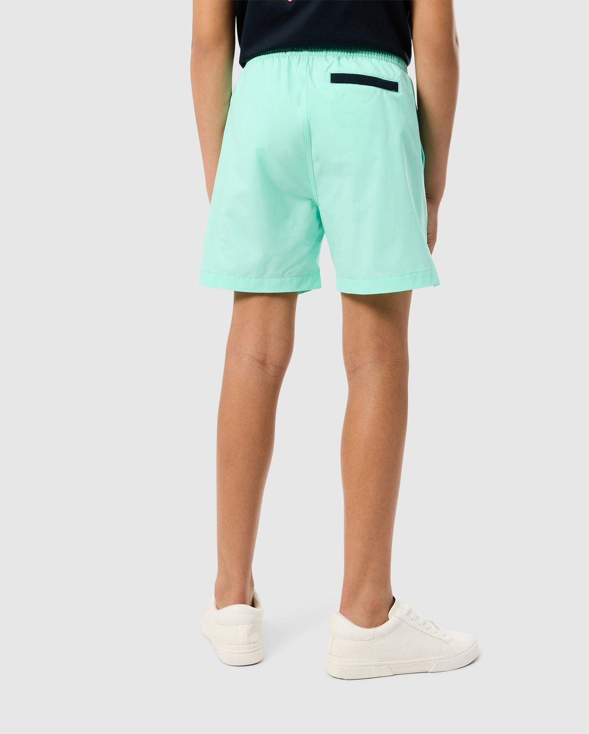 A person is standing facing away, showcasing the back view of their outfit. They are wearing Psycho Bunny's KIDS PARKER HYDROCHROMIC SWIM TRUNK - B0W646C200 in light green with a black pocket detail, a dark top, and white sneakers against a plain light grey background.