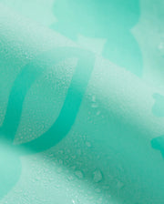 A close-up image of light turquoise fabric with water droplets. The fabric has a soft texture and a slight sheen, with parts of a darker turquoise pattern or design visible beneath the droplets, reminiscent of the KIDS PARKER HYDROCHROMIC SWIM TRUNK - B0W646C200 by Psycho Bunny reacting beautifully to moisture.
