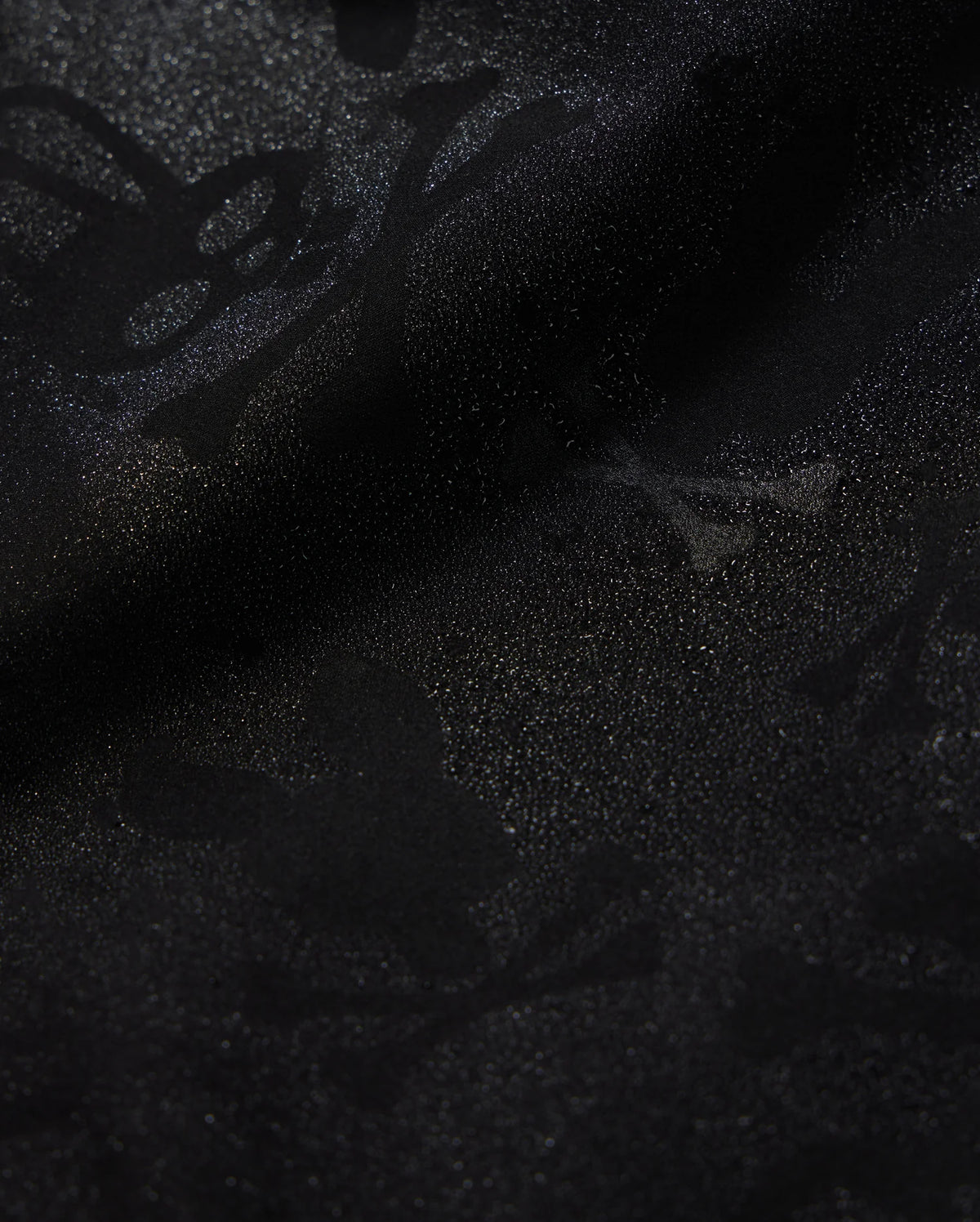 A detailed close-up of a Psycho Bunny KIDS PARKER HYDROCHROMIC SWIM TRUNK - B0W646C200 fabric displaying a subtle textured pattern with skull and crossbones and other abstract shapes. The slightly shiny, quick-dry material adds depth and dimension to the dark hydrochromatic pattern.