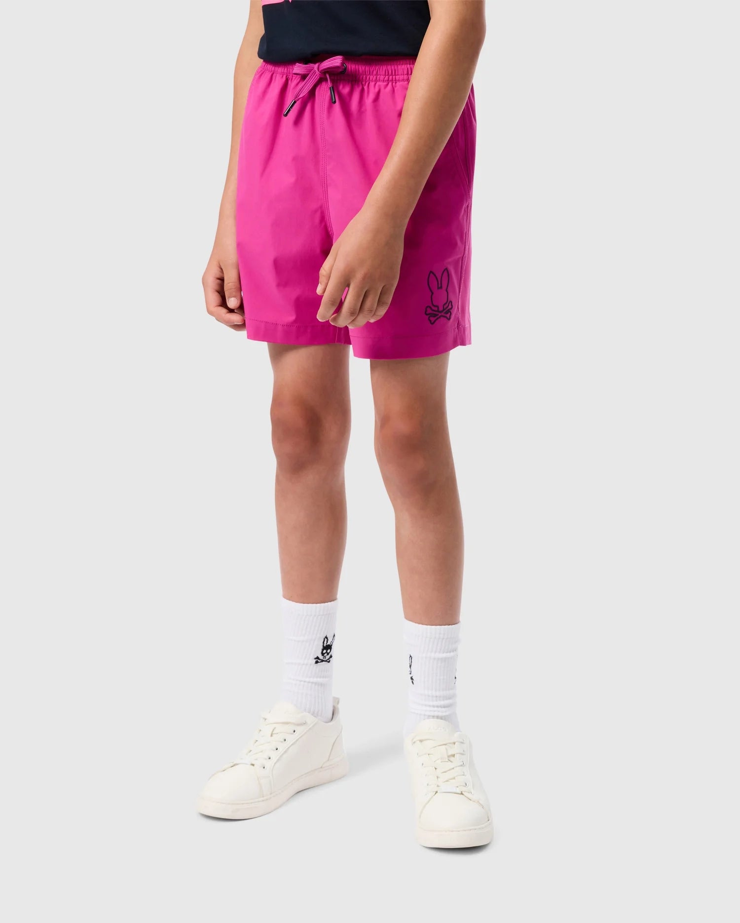 A person standing on a white background is wearing a black shirt, bright pink Kid Parker Hydrochromic Swim Trunk by Psycho Bunny featuring an embroidered bunny pattern on the left leg, white crew socks with a logo, and white sneakers. Only the torso and legs are visible.