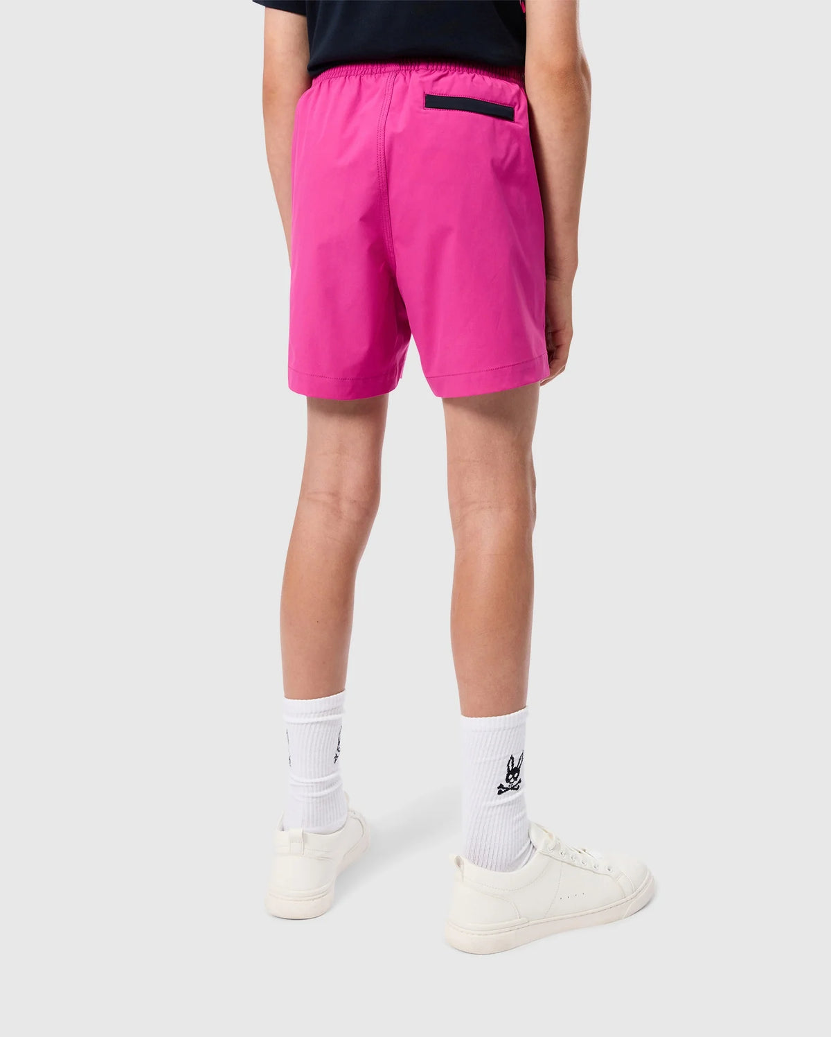 A person standing with their back to the camera, wearing bright pink Psycho Bunny KIDS PARKER HYDROCHROMIC SWIM TRUNKS (B0W646C200), a black shirt, white socks, and white sneakers. The shorts feature a black pocket on the right side. The background is plain and gray.