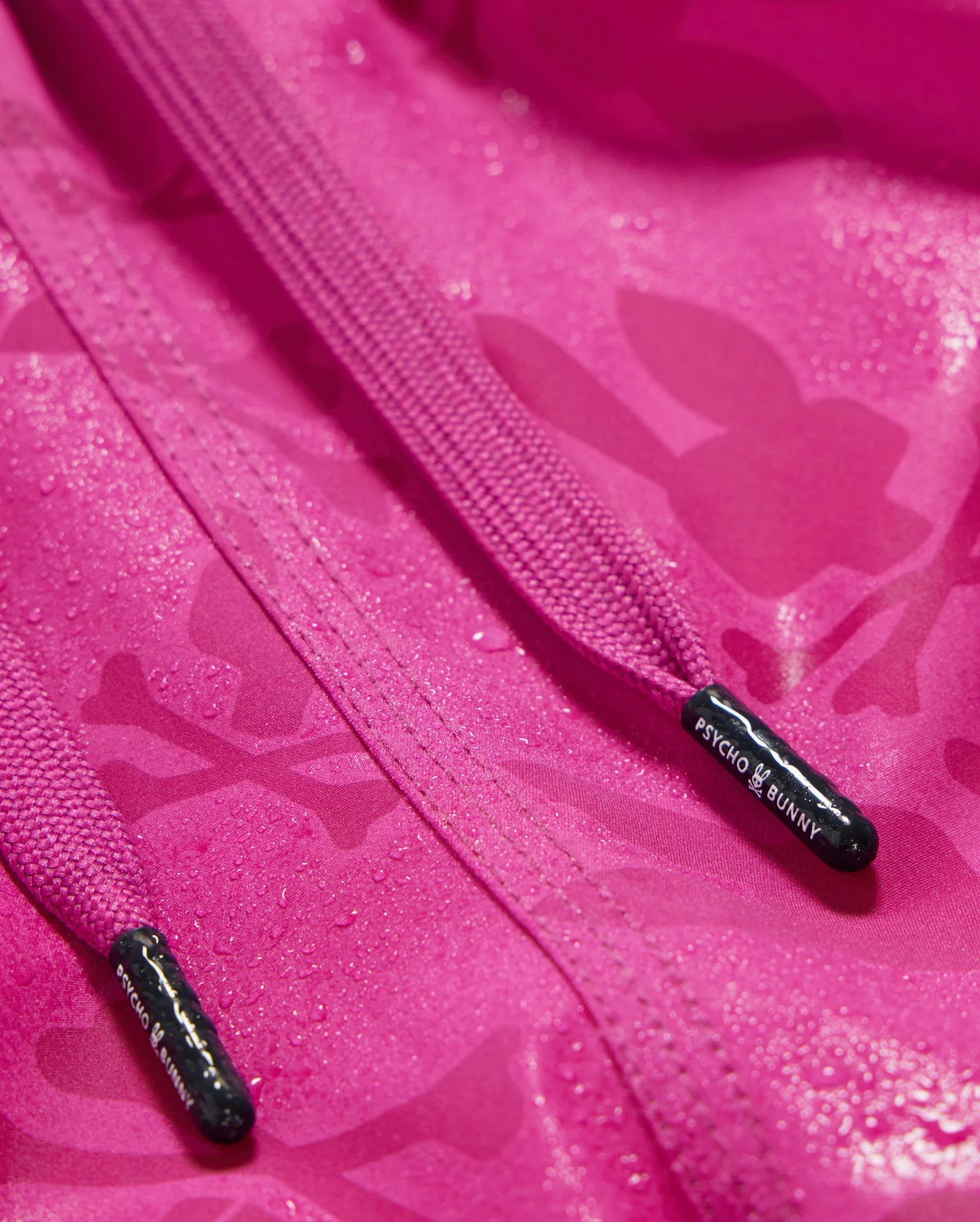 Close-up image of a pink KIDS PARKER HYDROCHROMIC SWIM TRUNK - B0W646C200 by Psycho Bunny, with water droplets on the fabric. The image prominently features two pink drawstrings, each with a black cap displaying the Psycho Bunny logo and branding in white text, alongside a subtle bunny pattern. The Psycho Bunny logo is also visible on the fabric.