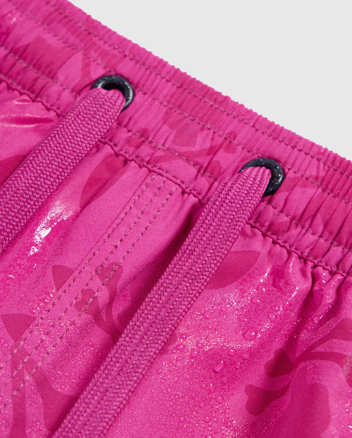 Close-up image of the KIDS PARKER HYDROCHROMIC SWIM TRUNK - B0W646C200 by Psycho Bunny, featuring a pair of bright pink swim trunks with hydrochromic fabric. The trunks have an elastic waistband and visible drawstrings. The fabric, adorned with a subtle bunny pattern, appears wet, reflecting light and showcasing its quick-drying properties.