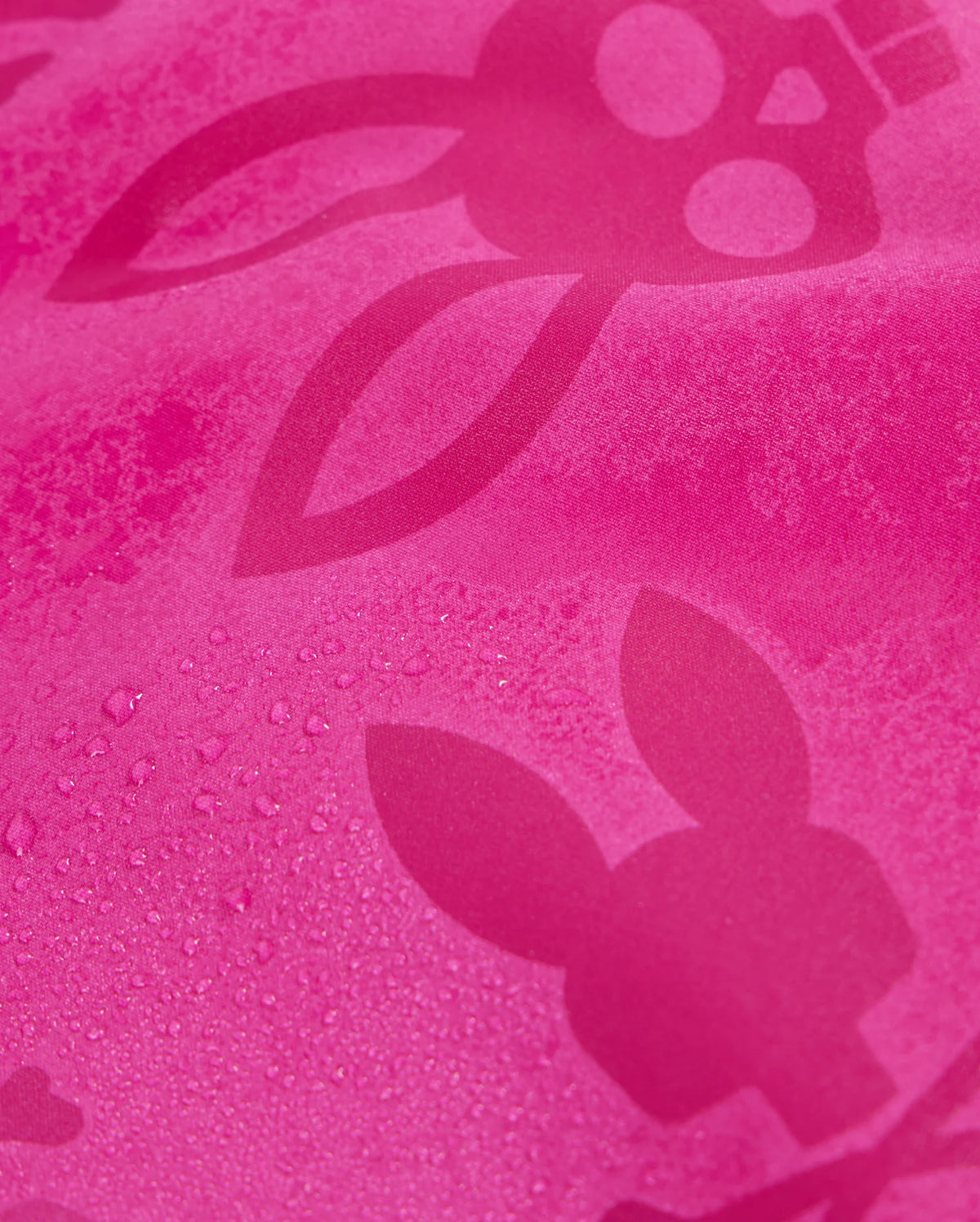 A close-up of the textured pink fabric of Psycho Bunny's KIDS PARKER HYDROCHROMIC SWIM TRUNK - B0W646C200, featuring a bunny pattern with long ears and skull-like faces. Some areas have visible water droplets, emphasizing the hydrochromic swim trunks' quick-drying properties.