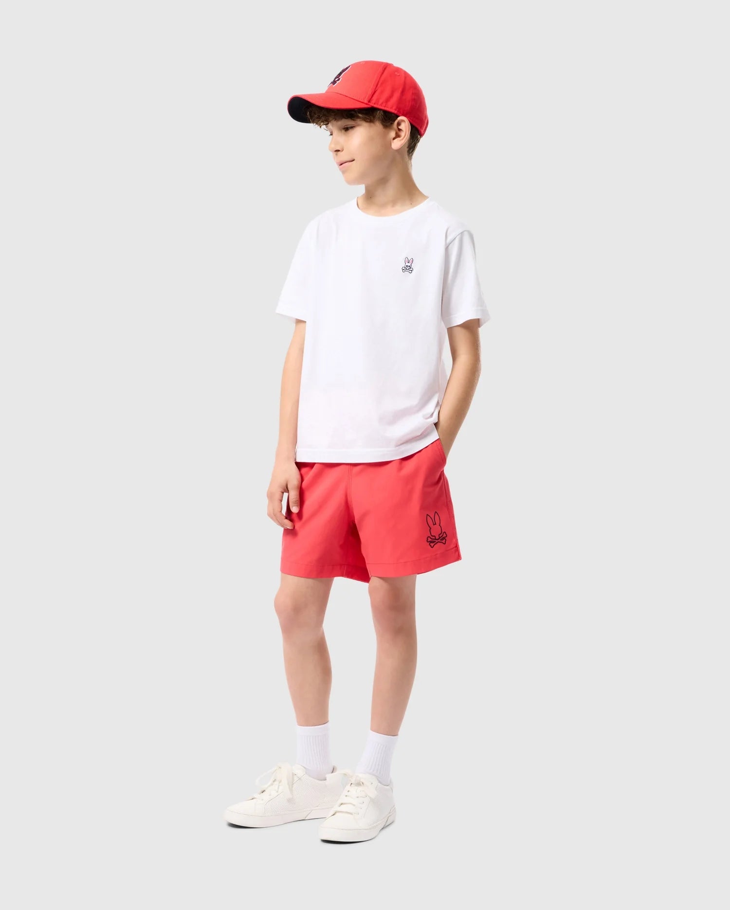 A young boy is standing against a plain background. He is wearing a red baseball cap, a white t-shirt with a small graphic on the chest, KIDS PARKER HYDROCHROMIC SWIM TRUNK - B0W646C200 by Psycho Bunny, white socks, and white sneakers. His hands are in his pockets, and he is looking to the side.