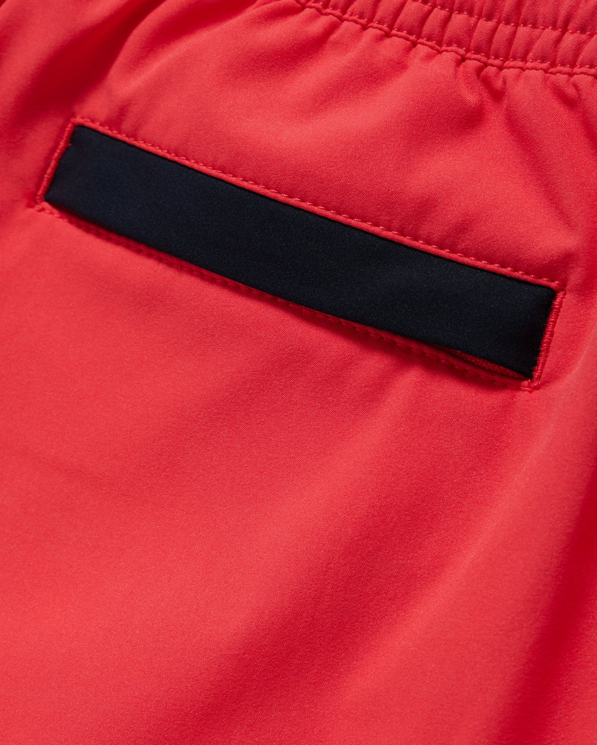 Close-up image of a back pocket on a piece of red hydrochromatic swim trunk fabric with an elastic waistband. The pocket has a black trim at the opening, contrasting with the bright red material. The quick-dry material appears smooth and slightly shiny. Featured product: KIDS PARKER HYDROCHROMIC SWIM TRUNK - B0W646C200 by Psycho Bunny.