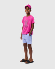 A smiling boy stands against a plain background wearing a pink cap, pink T-shirt with a bunny graphic, light blue Psycho Bunny KIDS MELVIN SWIM TRUNK - B0W665C200, and black slide sandals. He has one hand in his pocket and looks relaxed and cheerful.