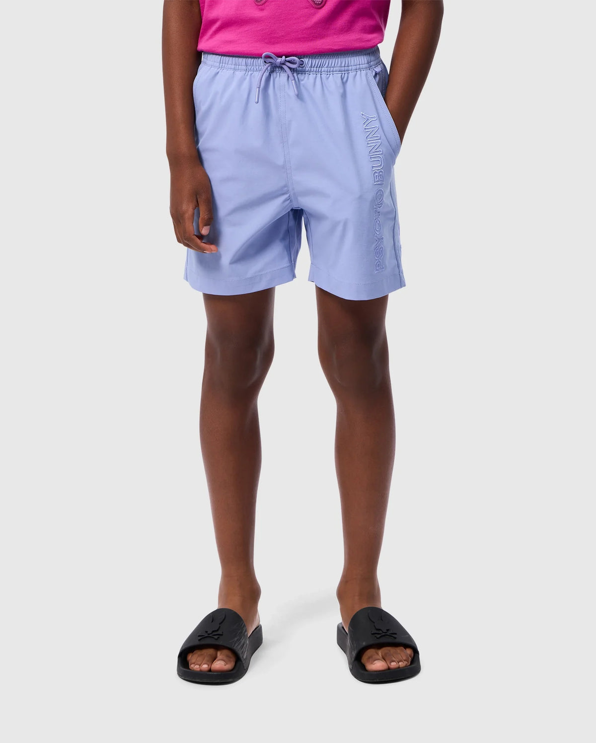 A person is wearing light purple KIDS MELVIN SWIM TRUNK - B0W665C200 by Psycho Bunny with a drawstring waist and the word "Savannah" printed on one leg. They are also dressed in a bright pink top and black slide sandals. The background is plain white.