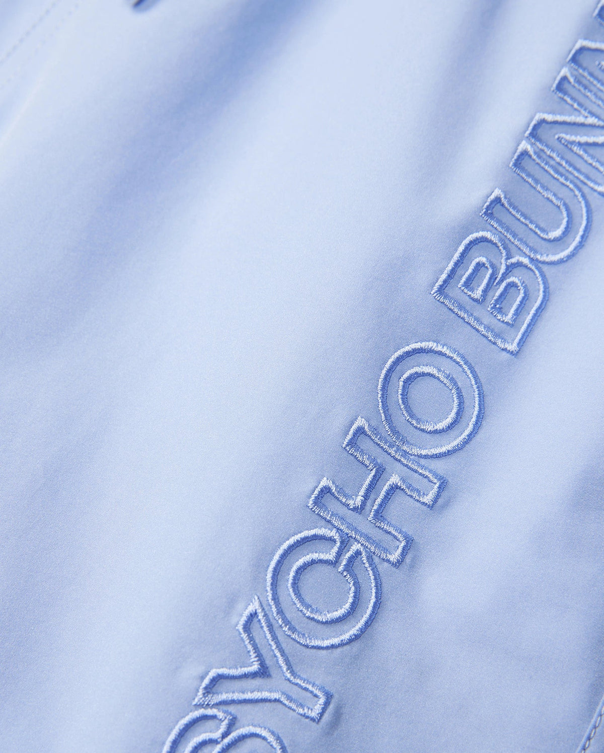 The product is a light blue KIDS MELVIN SWIM TRUNK (B0W665C200) by Psycho Bunny, featuring "PSYCHO BUN" partially embroidered in blue thread. This quick-dry fabric ensures comfort and style by the water.