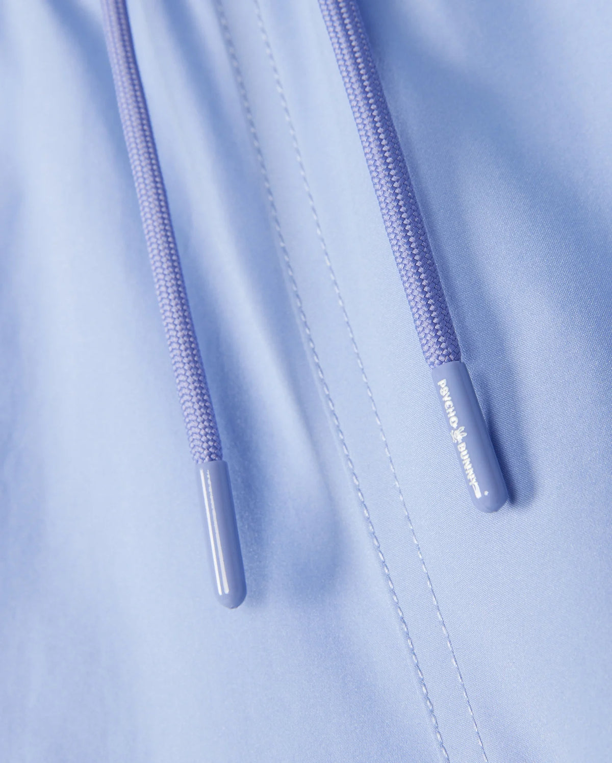 Close-up of the Psycho Bunny KIDS MELVIN SWIM TRUNK fabric in light blue, with lavender drawstrings featuring branded text on the tips. The quick-dry material appears smooth and slightly shiny with visible stitching.