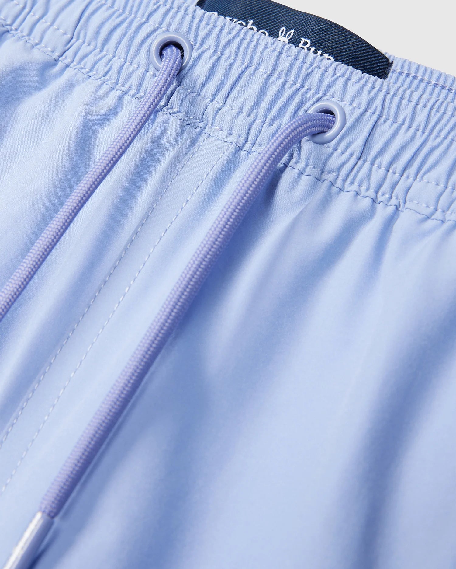 A close-up of Psycho Bunny's KIDS MELVIN SWIM TRUNK - B0W665C200 in light blue shows an elastic waistband with matching drawstrings. A small black label and embroidered logo are visible, suggesting a smooth, quick-dry finish likely from lightweight fabric.