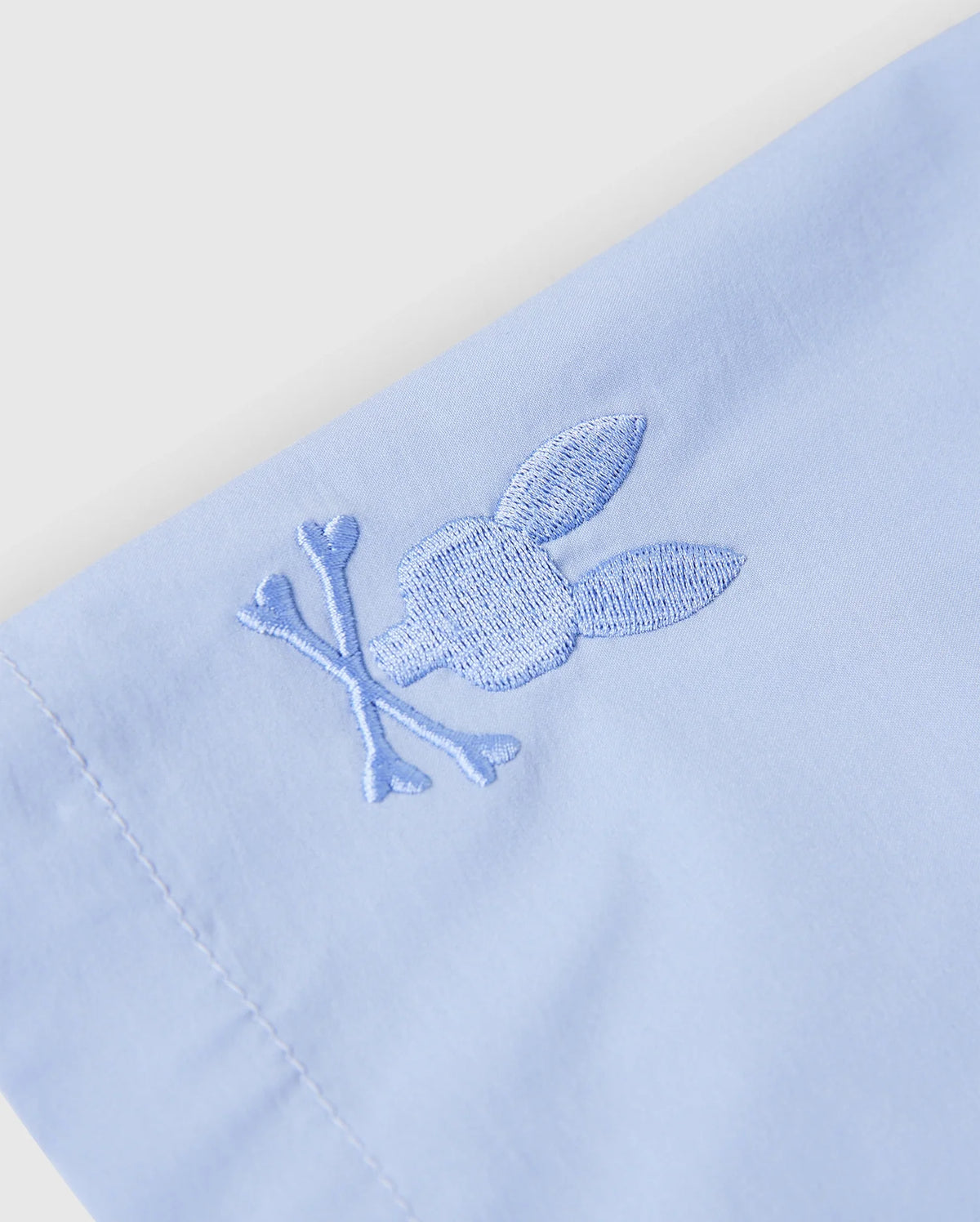 Close-up of the KIDS MELVIN SWIM TRUNK by Psycho Bunny in light blue, showcasing an embroidered rabbit head and crossbones in matching thread. The quick-dry fabric's stitching is detailed, smooth, and well-tailored. Model: B0W665C200.