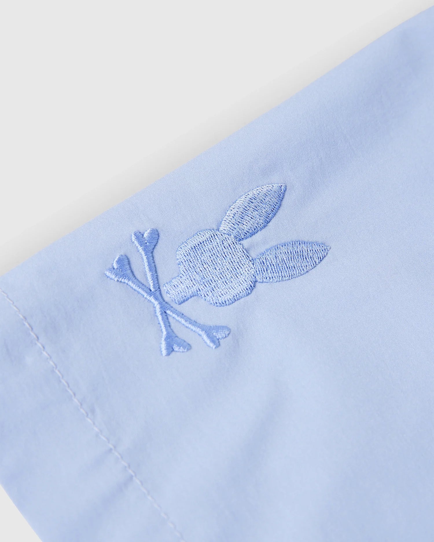 Close-up of the KIDS MELVIN SWIM TRUNK by Psycho Bunny in light blue, showcasing an embroidered rabbit head and crossbones in matching thread. The quick-dry fabric's stitching is detailed, smooth, and well-tailored. Model: B0W665C200.