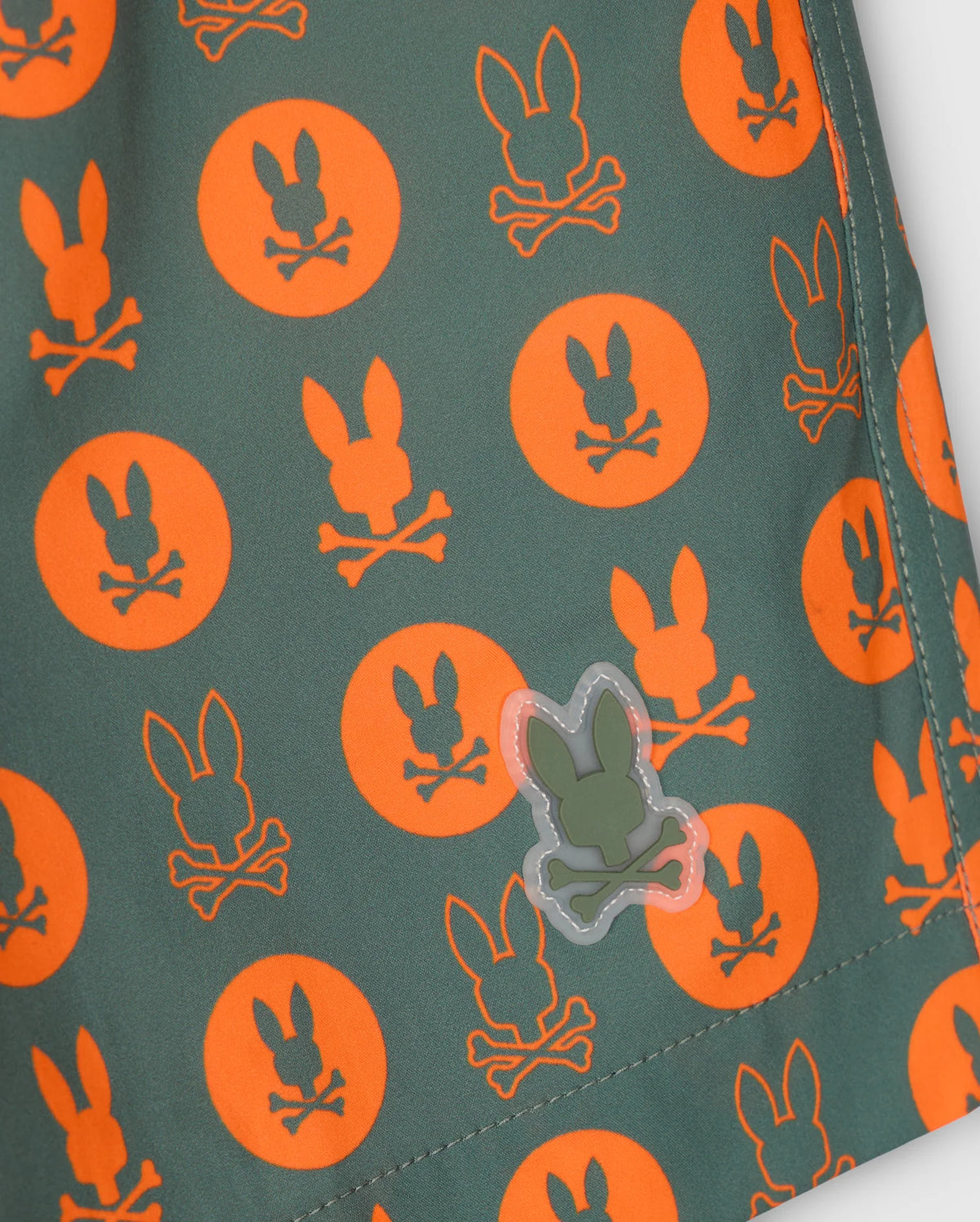 Close-up of the KIDS ARNOLD PRINT SWIM TRUNK - B0W820D200 fabric, showcasing orange bunnies and crossbones in circles against a green backdrop. A small green bunny silhouette adds a unique touch to the pattern, crafted with quick-dry material for enhanced comfort and functionality by Psycho Bunny.