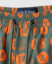 Close-up of the KIDS ARNOLD PRINT SWIM TRUNK - B0W820D200 featuring green swim trunks with orange patterns, including a bunny and crossed bones. The quick-dry material ensures comfort, complemented by a waistband adorned with a black Psycho Bunny label. These trunks are designed with stretch mesh lining and green drawstrings for the perfect fit.