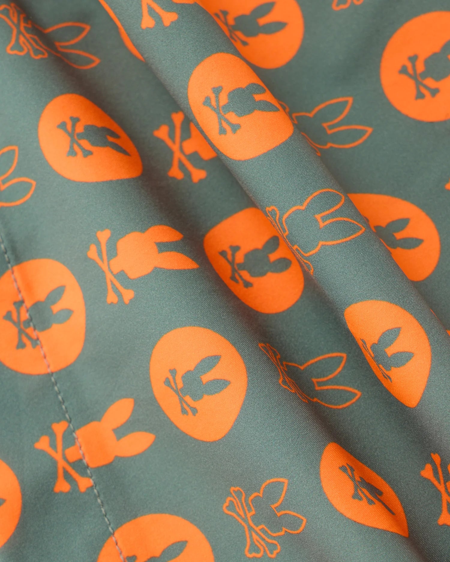 The KIDS ARNOLD PRINT SWIM TRUNK - B0W820D200 by Psycho Bunny is designed with quick-dry fabric featuring an orange print on a dark green backdrop adorned with circles containing silhouettes of bunnies and crossed bones. The pattern repeats seamlessly, complemented by a fun stretch mesh lining.