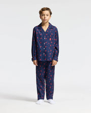 A child stands dressed in the KIDS ALL OVER PRINT FLANNEL PAJAMA SET - B0X356E200 from Psycho Bunny, showcasing its soft fabric and navy blue and red patterns. This lightweight sleepwear includes a button-up top paired with matching pants. The child, with brown hair and bare feet, is set against a simple white background.