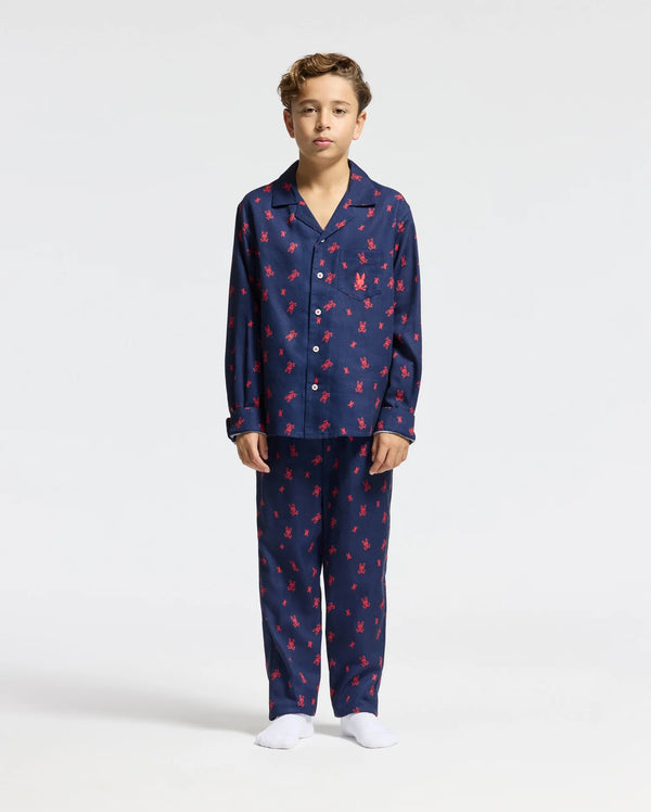 A child stands dressed in the KIDS ALL OVER PRINT FLANNEL PAJAMA SET - B0X356E200 from Psycho Bunny, showcasing its soft fabric and navy blue and red patterns. This lightweight sleepwear includes a button-up top paired with matching pants. The child, with brown hair and bare feet, is set against a simple white background.